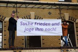 Bischof Thomas Adomeit (li.) und Birgit Carmona Scheider vom Bischofsbüro befestigen das Banner mit den Psalmworten „Suche Frieden und jage ihm nach!“ (Psalm 34,15) am Gebäude des Oberkirchenrates im Oldenburger Philosophenweg. Foto: ELKiO/D.-M. Grötzsch